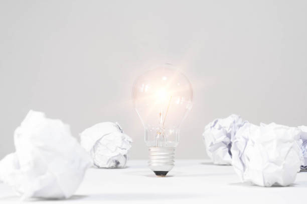 Glowing light bulb surrounded by crumpled paper balls on a white background, symbolizing creativity and brainstorming.