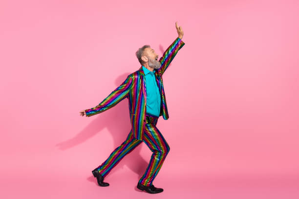 istock Eccentric senior man in a vibrant patterned suit dancing enthusiastically on a pink background, embodying fun, fashion, and charisma 2195235253