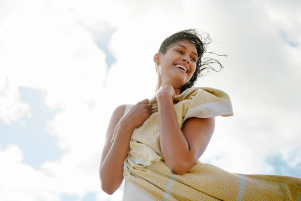 A cheerful woman encapsulates freedom while standing outdoors with a bright smile, wrapped in a towel against a sky filled with light clouds.
