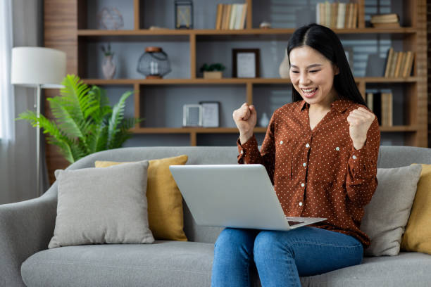 istock Asian woman celebrating success while using laptop at home, expressing achievement and excitement on comfortable sofa, demonstrating happiness and accomplishment in cozy living room setting 2195236553