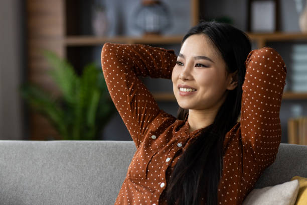 istock Content asian woman in polka dot blouse relaxing on sofa in modern home setting, smiling with hands behind head, expressing relaxation and happiness in a cozy environment. 2195236568