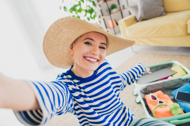 istock Joyful young woman taking selfie while packing suitcase for a vacation at home in a striped pullover 2195236848