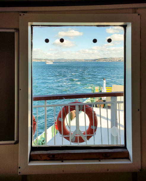 Waves gently lap against the bow of the ship as a bell hangs above, offering a view of the city skyline along the Bosphorus strait on a cloudy day.
