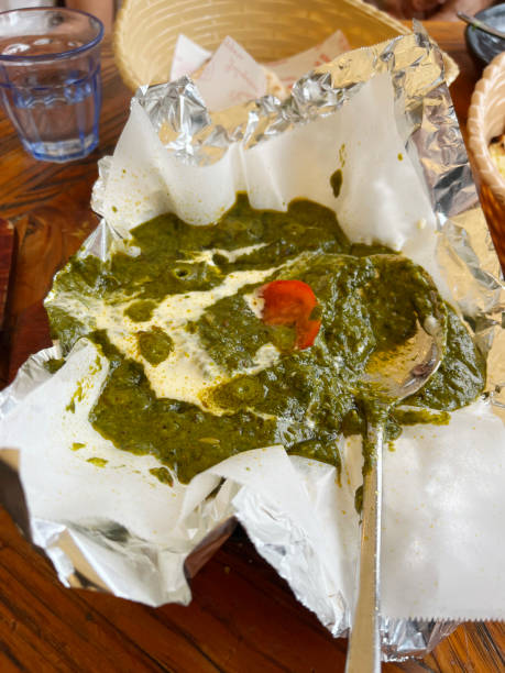 A rustic serving of palak paneer curry bubbling in a pan, surrounded by cooking paper on a wooden table, with garlic naan visible in the background