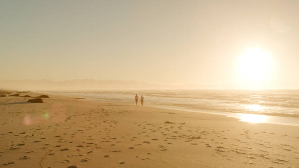 istock Two Silhouetted Figures Walk Along Beach By Waves At Sunset 2195260452
