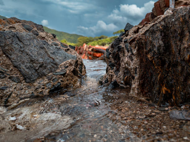 A small stream flows through rugged rocky formations in Tayrona National Park, Colombia. The earthy textures of the rocks contrast with the clear water