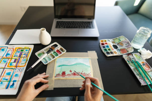 Woman is sitting at a desk in her living room and is watching tutorials on her laptop to learn how to paint with water colors,
