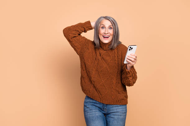 istock Stylish elderly woman joyfully celebrating an accomplishment while using a laptop, standing against a plain beige background 2195266073