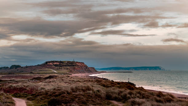 A scenic view of Southbourne beach with cliffs, dunes, and coastal paths under a cloudy sky. The tranquil sea and distant horizon create a peaceful and picturesque coastal landscape.