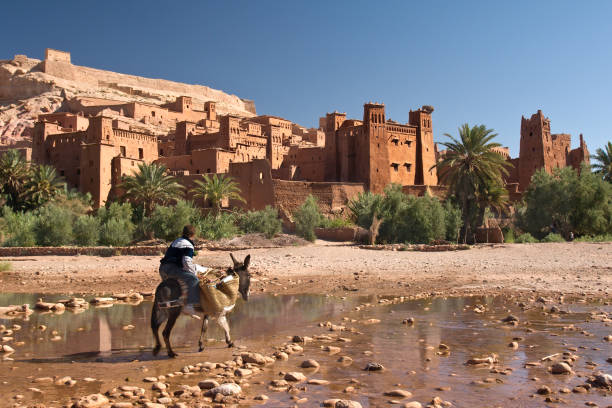 Ouarzazate, Morocco - June 24, 2010: A man rides a donkey across shallow water heading toward Ait Benhaddou Kasbah; the  most famous kasbah in Morocco which is a movie set & UNESCO site.
