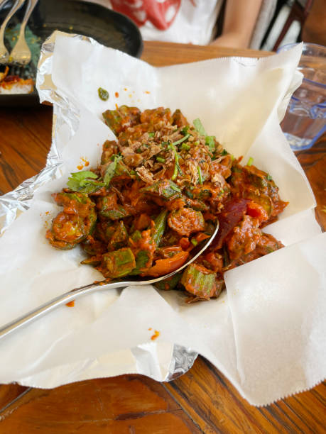 A rustic serving of Okra curry bubbling in a pan, surrounded by cooking paper on a wooden table, with garlic naan visible in the background
