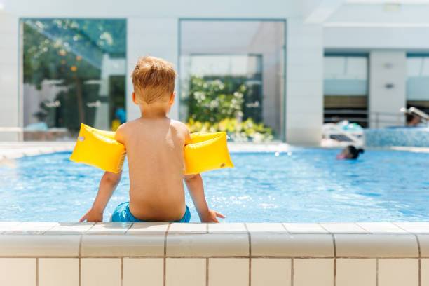 istock Back view cute adorable little caucasian blond boy kid wear inflatable armband sit poolside indoor pool edge before swimming hotel spa thermal resort. Happy child enjoy summer vacation travel family 2195284082
