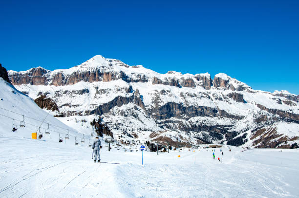 Arabba, Italy - March 24, 2024: Easy ski slope with skiers and chairlift ski lift in Arabba ski resort in Dolomites, Italy. Sella Group mountain massif in the background. Blues sky with copy space