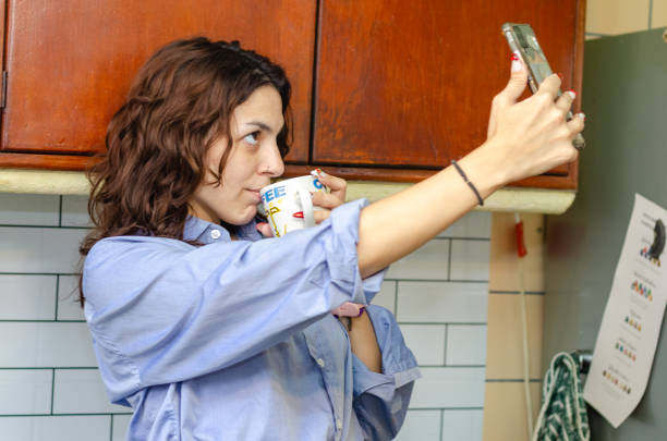 Young female employee or student taking a selfie with her mobile phone while drinking a coffee in the kitchen
