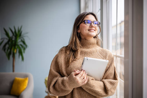 istock Woman in cozy sweater smiling while holding tablet near window in modern interior space during daytime 2195306047