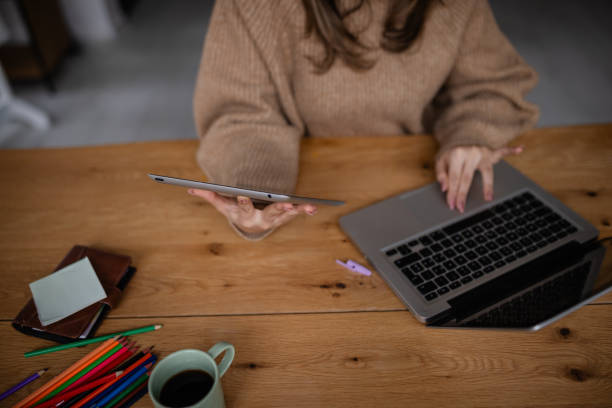 A young adult woman sits at a wooden table in a contemporary workspace, multitasking with a laptop and tablet. Colorful stationery and a coffee cup add a vibrant touch to the setting.