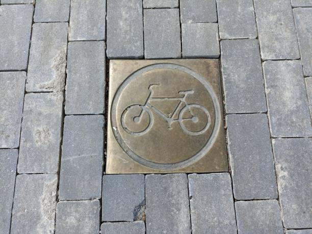 A section of pavement paved with grey rectangular tiles. In the centre is a square metal plate with a picture of a bicycle enclosed in a circle. This may indicate a bicycle path or a place where bicycles are allowed.