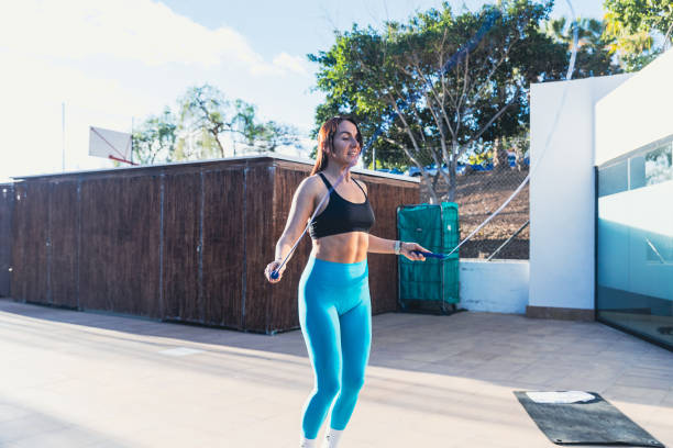A woman is jumping rope in a park. The sky is blue and the sun is shining. The woman is smiling and seems to be enjoying herself