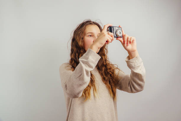 A young girl with long wavy hair, wearing a beige sweater, holding a camera up as if taking a picture. She is focused and positioned in a well-lit studio environment against a plain white background, exuding creativity and intent