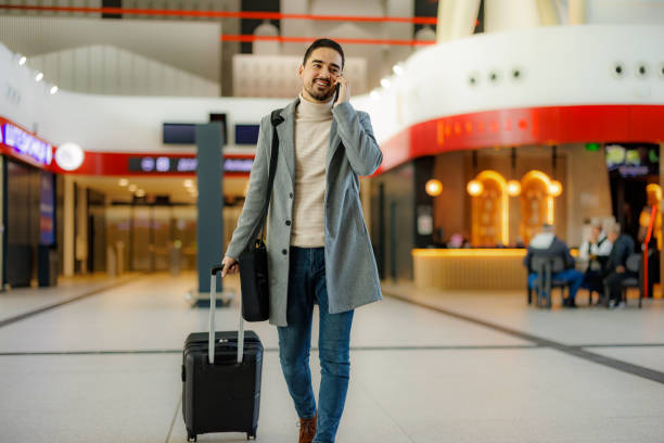 Businessman walking in airport terminal while talking on phone and pulling suitcase