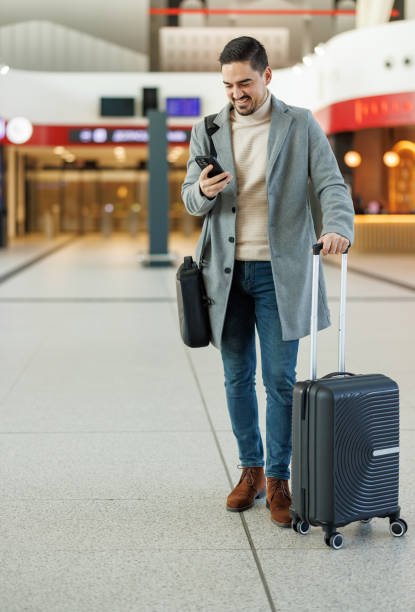 istock Businessman walking in airport terminal using smartphone and pulling suitcase 2195330980