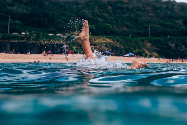Captivating low-angle shot of carefree legs kicking up water, framed by vibrant blue ocean waves and a lively sandy beach. Lush green cliffs and beachgoers with umbrellas in the background complete this tropical paradise scene. Perfect for themes of summer, vacation, relaxation, joy, and outdoor fun.