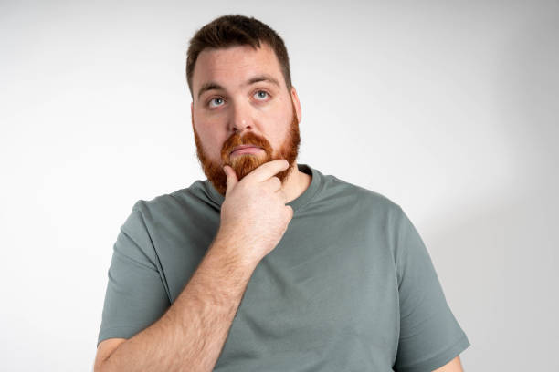 istock A young bearded man stands thoughtfully with his hand on his chin against a grey background. 2195337917