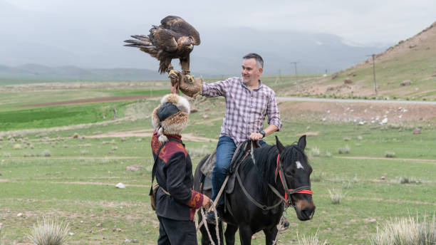 Issyk Kul, Kyrgyzstan - 23 May 2022: Eagle trainer assisting a tourist to hold an golden eagle, an experience vacation