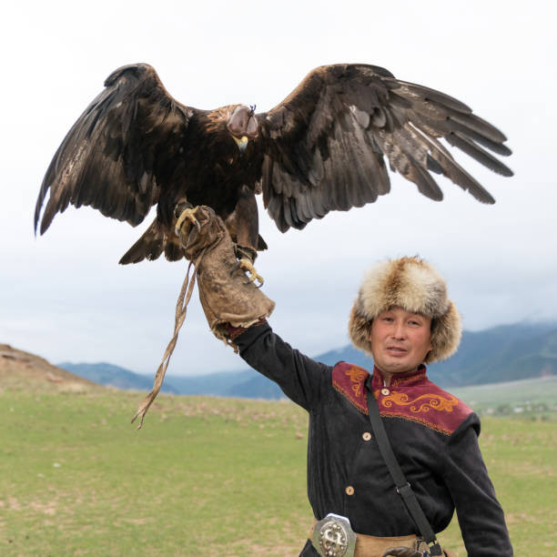smiling chinese female tourist takes a selfie with a Kazakh eagle hunter dressed in traditional fur clothing, holding a golden eagle against a rugged mountain backdrop in Mongolia.