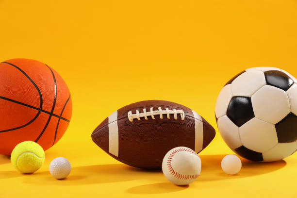 A closeup image of a stack of sports balls against a reflective blue background. In the stack is a football, baseball, basketball and tennis ball.