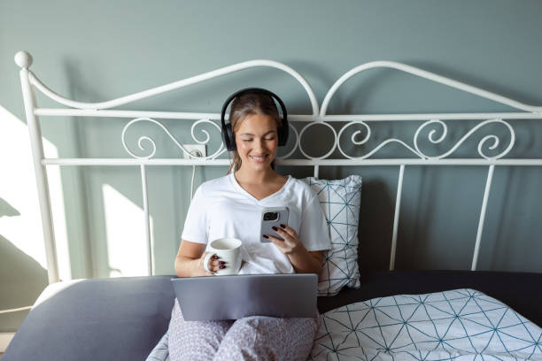 Beautiful woman with coffee cup using headphones and consuming Internet content via phone and laptop in bedroom