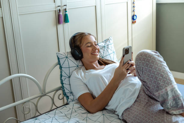 istock Young woman using mobile phone and wireless headphones while lying comfortably in bed 2195353370