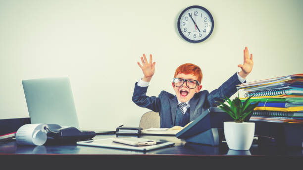 istock Humorous image of happy enthusiastic young boy working on a laptop computer at a desk. He is wearing a suit and is celebrating with his arms in the air. 2195358849