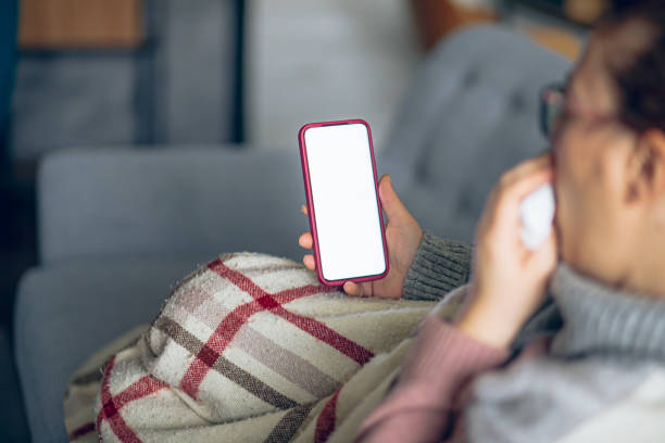 istock Sick woman sits on the sofa and wipes her nose with a handkerchief while using mobile phone 2195359159