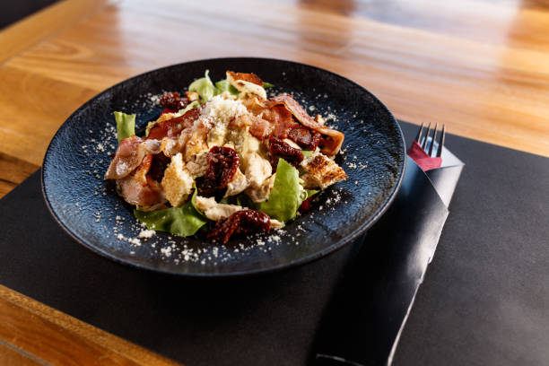 A close-up view of a Caesar Salad featuring crispy bacon, sun-dried tomatoes, and croutons. Served on a black plate, it highlights fresh ingredients for a savory and delicious dining experience