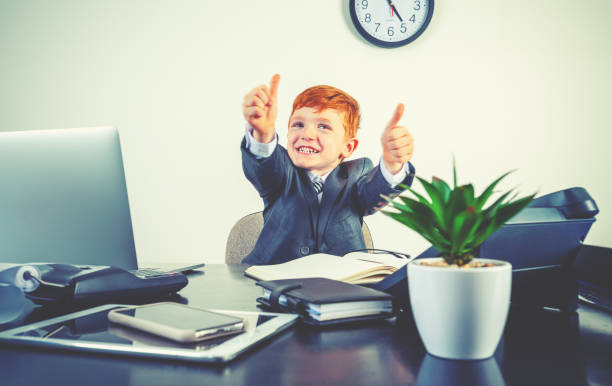 istock Humorous image of happy enthusiastic young boy working on a laptop computer at a desk. He is wearing a suit and is giving a positive thumbs up 2195361438