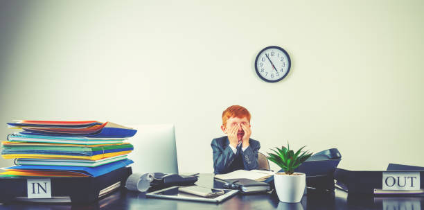 istock Humorous image of stressed and upset young boy working on a laptop computer at a desk. He is on the phone wearing a suit and is looking overworked 2195361453