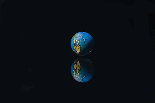 A small blue glass globe with gold details reflected on a black surface. The background is also black.