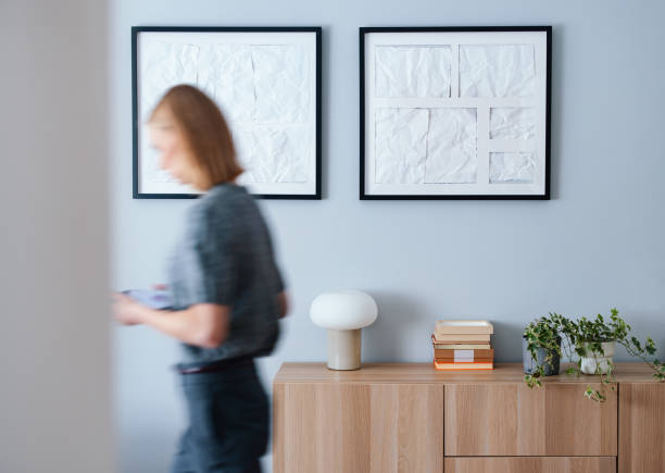 A modern interior featuring framed wall art, a wooden cabinet with decor items, and a blurred woman. The scene exudes simplicity, elegance, and an artistic touch, ideal for design or lifestyle concepts.