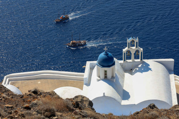 A whitewashed church with a vibrant blue dome and three traditional bells overlooks the vast Aegean Sea. Two wooden sailing boats glide gracefully across the deep blue waters under the bright midday sun. The scene radiates tranquility and timeless charm, with warm light highlighting the simplicity and elegance of Cycladic architecture. A gentle breeze and clear skies complete this serene Mediterranean moment