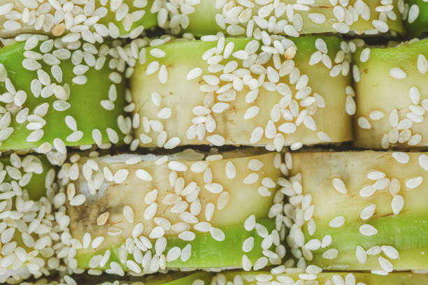 A close-up image of a plate of sushi rolls topped with white sesame seeds. The rolls feature a green filling, likely avocado, and a white filling, possibly imitation crab or tofu