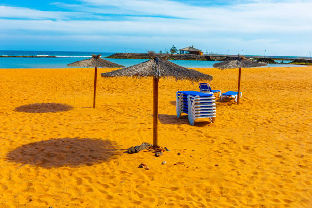 Beach scene with straw umbrellas casting shadows on the sand and stack of blue and white lounge chairs. Background with calm ocean with breakwater suggesting a tranquil and inviting seaside location