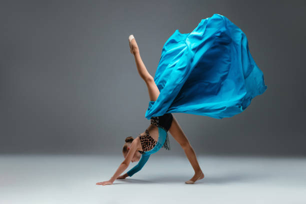 istock Graceful ballerina dancing on grey studio background. Cheerleader dancing with flying blue cloth fabric. Dancer in sport leotard doing Split. Child, artist, movement, action and motion concept 2195391239