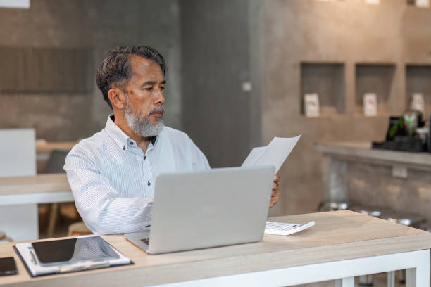 istock Diligent male businessman examining paperwork while working at a desk with laptop in a modern office 2195392863