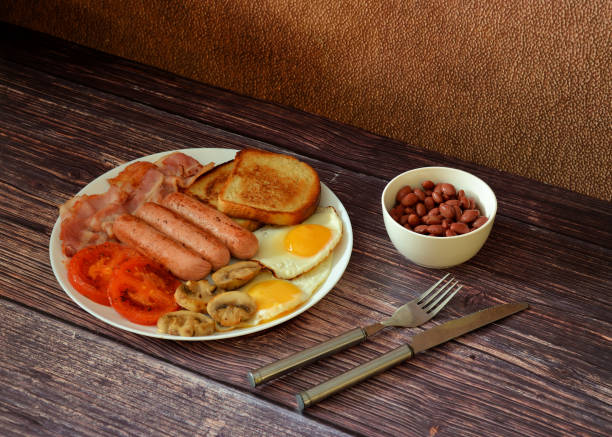A hearty and nutritious breakfast, a plate with fried sausages, bacon, scrambled eggs, tomatoes, mushrooms and wheat croutons on a wooden table, next to a cup of beans and cutlery. Close-up.