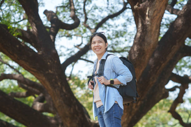 istock An Asian woman wearing a denim jacket and backpack smiles under a large tree in a forest. 2195406712