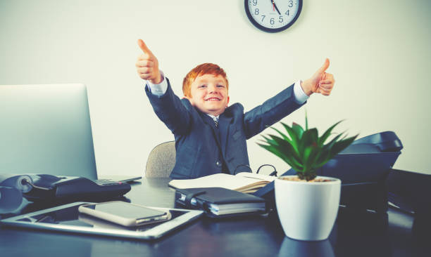 istock Humorous image of happy enthusiastic young boy working on a laptop computer at a desk. He is wearing a suit and is giving a positive thumbs up 2195410729