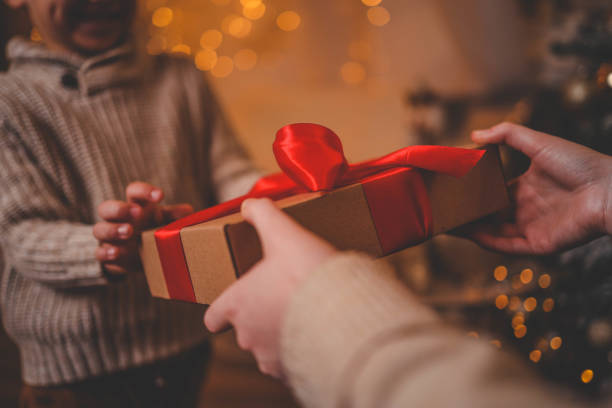 Hands exchanging a festive gift box with a red ribbon under a beautifully decorated Christmas tree. Cozy holiday setting with candles, miniature houses, and a warm atmosphere