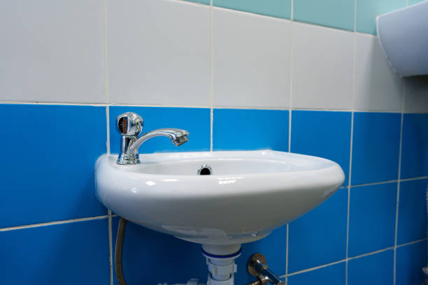 A compact white sink installed against a blue-tiled wall features a shiny chrome faucet. Natural light illuminates this clean bathroom area, highlighting the tiles and sink design.