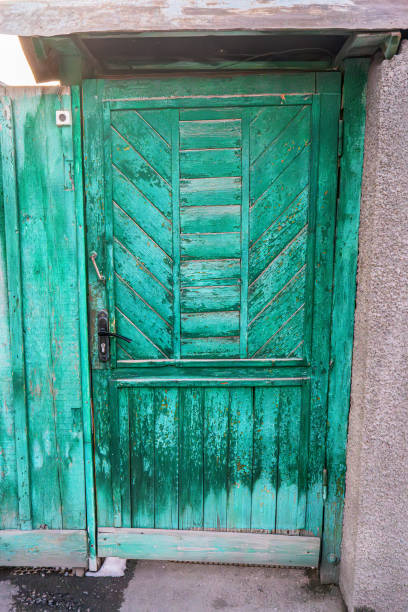 A rustic green wooden door with peeling paint and a textured, weathered surface. The door features diagonal planks and an old metal handle, framed by a concrete wall, creating a charming, vintage look.